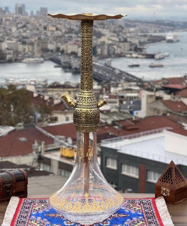 Turkse traditionele waterpijp met gegraveerde messing schacht en decoratieve vaas, geplaatst op een kleurrijk tapijt met uitzicht over Istanbul.