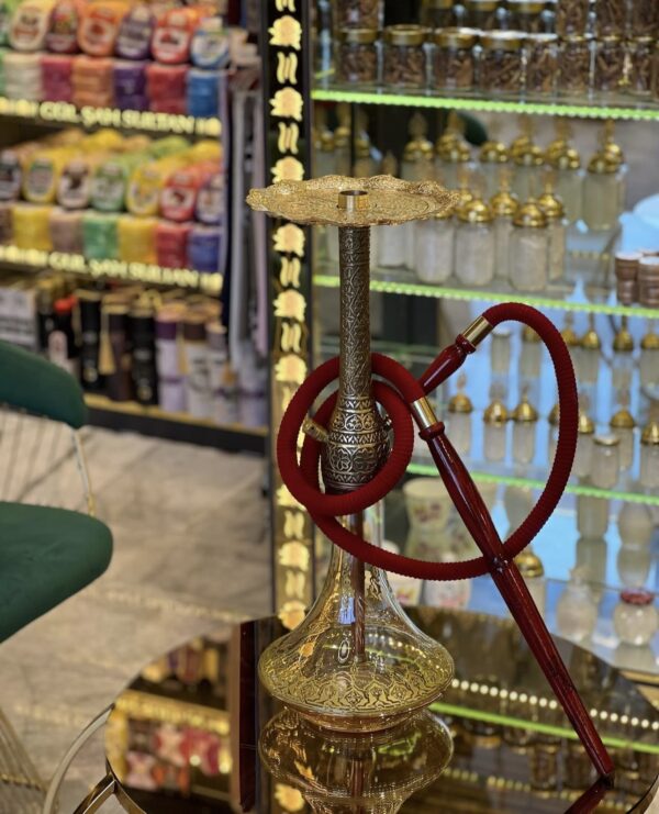 Luxe Turkse waterpijp met gegraveerde gouden vaas en messing schacht, voorzien van een rode slang, gepresenteerd op tafel in een oosterse winkel.